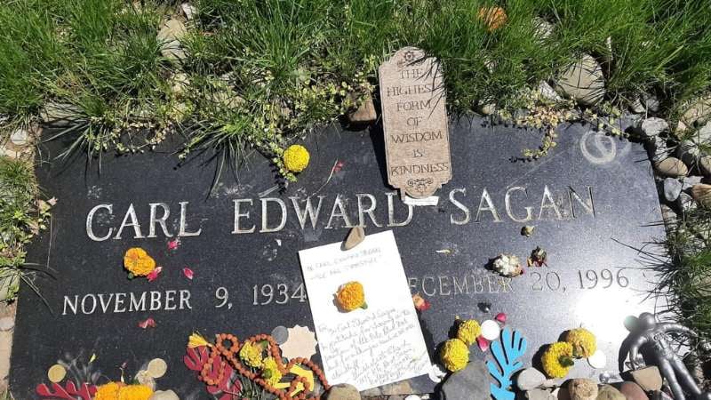 Carl Sagan’s gravestone with a plaque reading “The highest form of wisdom is kindness,” surrounded by flowers and tributes.